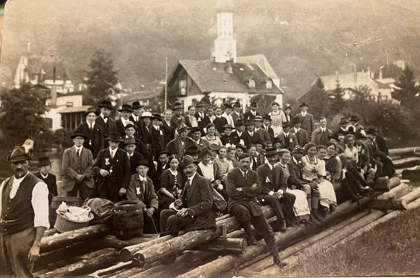 Historisches Schwarzweiß-Foto eines Vergnügungsfloßes mit vielen Fahrgästen in festlicher Kleidung bei Wolfratshausen, 1920er/1930er Jahre, im Hintergrund Häuser und ein Kirchturm